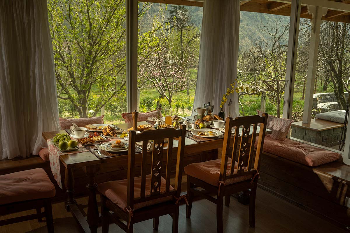 A dining table at the homestay with breakfast dishes like eggs, home-baked bread, jams and preserves facing big windows with a view of lush verandah