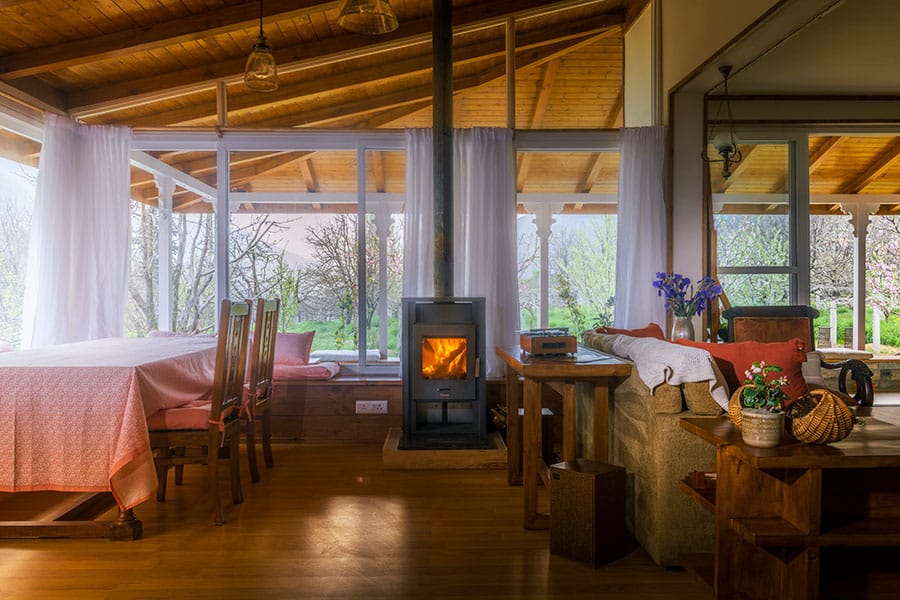 A rustic drawing room with wooden flooring, metal fireplace, dining table, sofa and big windows facing the orchard